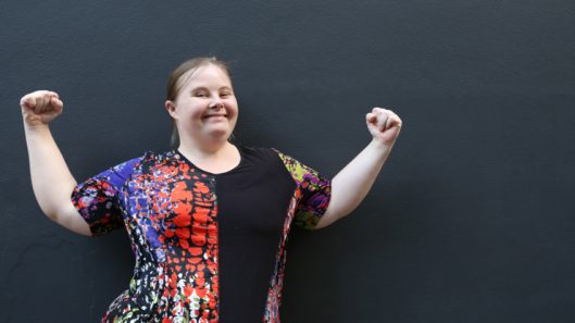 A woman with Down Syndrome holds her arms up in a show of strength.
