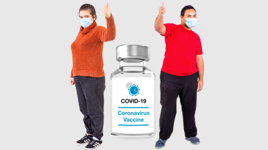 Two people standing next to a giant COVID vaccine and giving a thumbs up sign.