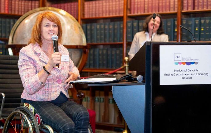 A woman with red hair using a wheelchair speaks into a microphone next to a lectern. Fiona McKenzie smiles in the background.