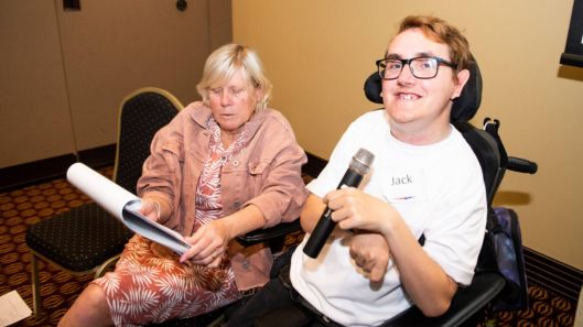 A man using a wheelchair holds a microphone and smiles. His support person holds a clipboard with notes.