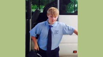 Finlay Browne, a young man with Down Syndrome, stands in his school uniform in front of an open bus door.