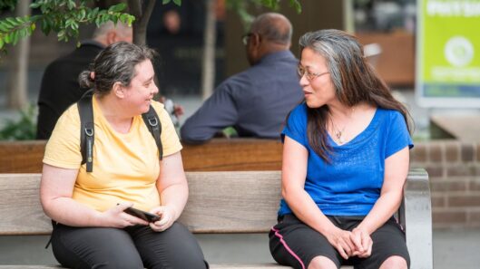 Two women with intellectual disability sit on a bench and have a conversation.