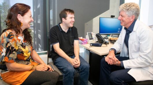 A man with intellectual disability sits in a doctor's office with a supporter and a doctor. They are all smiling.