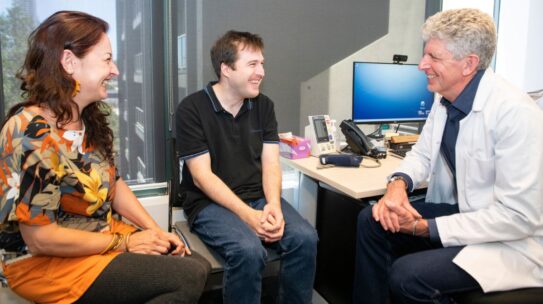 A man with intellectual disability sits in a doctor's office with a supporter and a doctor. They are all smiling.