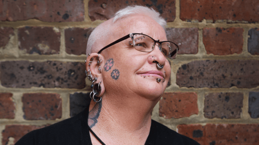 An intellectual disability advocate with tattoos and piercings smiles and looks away from the camera.