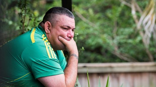 A First Nations man with intellectual disability stands on a balcony. He has his hand over his mouth and he looks worried.